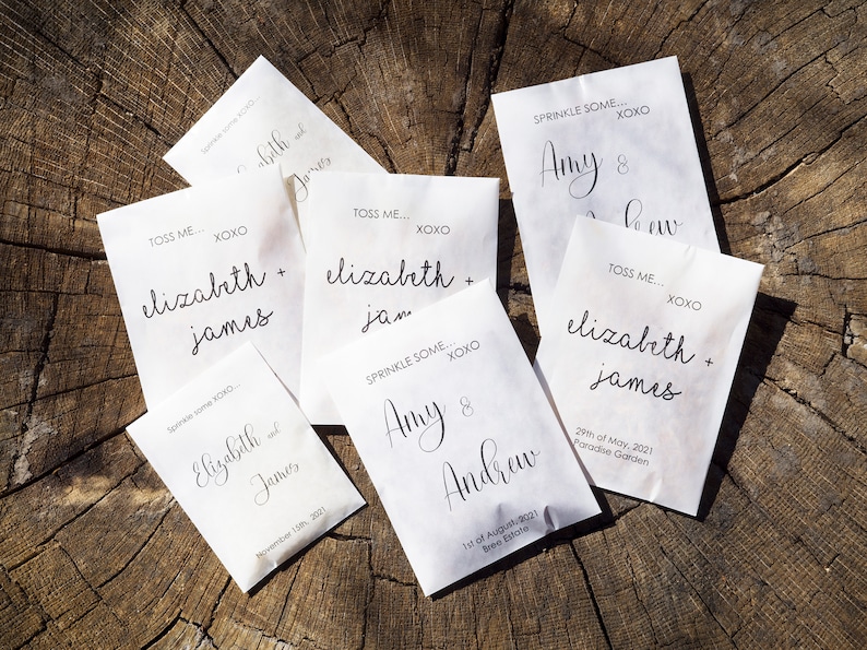 May include: Six white paper envelopes with black text. The envelopes are for wedding favors and have the names of the couples and the date of their wedding. The text on the envelopes reads: "Toss Me... XOXO Elizabeth + James", "Sprinkle Some... XOXO Elizabeth + James", "Toss Me... XOXO Elizabeth + James", "Sprinkle Some... XOXO Amy & Andrew", "Sprinkle Some... XOXO Amy & Andrew", and "Toss Me... XOXO Elizabeth + James".