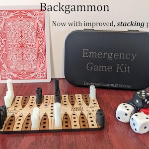 May include: A black and brown backgammon game set with white and black playing pieces. The game board is in a wooden case with the words "Emergency Game Kit" and "DAVESTARGAMING.ETSY.COM" printed on it. The game set also includes dice and a playing card for scale.