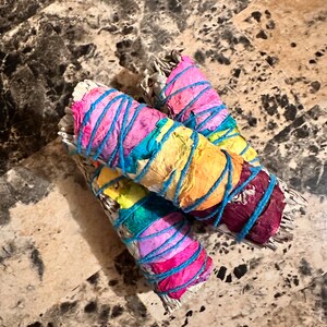 May include: Three white sage smudge sticks wrapped in colorful paper and tied with blue string. The paper is a rainbow of colors, including pink, yellow, green, and blue.