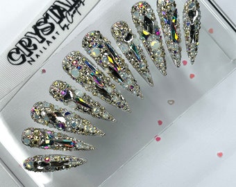 Peut inclure: Un ensemble de dix faux ongles en argent en forme de stiletto avec un design en cristal. Les ongles sont décorés de strass transparents, irisés et blancs.