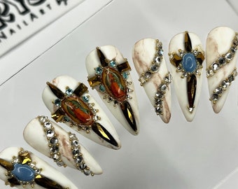 May include: White press-on nails with a marble design and gold accents. Each nail features a small cross with a religious image and rhinestones.