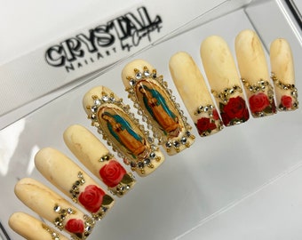 May include: Set of long, beige press-on nails with a marble effect. Each nail is decorated with gold accents, rhinestones, and a religious image of the Virgin Mary. Some nails also feature red roses.