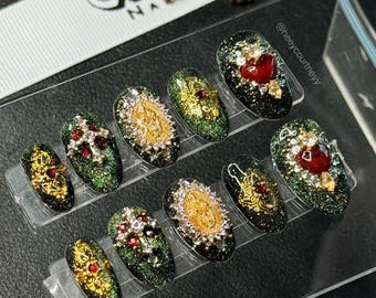 May include: A set of press-on nails with a glittery green and black base. Each nail features ornate gold and silver embellishments, including crosses, hearts, and religious iconography. The nails are displayed on a clear plastic holder.