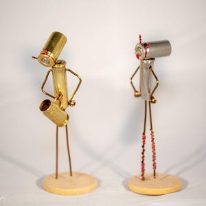 May include: Two small metal figures made from recycled bullet casings. The figure on the left is holding a saxophone and is made from gold-colored casings. The figure on the right is made from silver-colored casings and is standing with its arms crossed. Both figures are standing on small wooden bases.