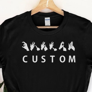 May include: Black t-shirt with white text and hand gestures spelling "CUSTOM" in British Sign Language. The shirt is hanging on a wooden hanger against a white background. A sprig of greenery is in the lower left corner.