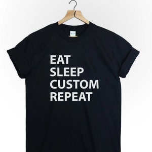 May include: Black t-shirt with the white text "EAT SLEEP CUSTOM REPEAT". The t-shirt is displayed on a wooden hanger against a white backdrop. The t-shirt has a crew neck.