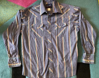 Può includere: Una camicia blu Wrangler con bottoni e strisce verticali arancioni, verdi e gialle. La camicia a maniche lunghe ha due tasche sul petto, un colletto e polsini con bottoni. Il logo Wrangler è visibile sul colletto.