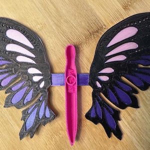 May include: A decorative butterfly wing set in black, purple, and pink. The wings are connected by a pink center piece and a purple band. The wings are detailed with a layered design, and the set is displayed on a wooden surface.