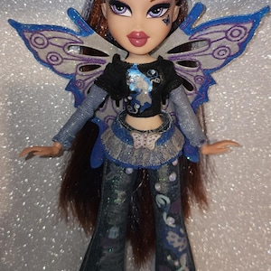 Puede incluir: Una muñeca Bratz con cabello castaño largo y un atuendo con temática de mariposas. La muñeca lleva una blusa negra con un diseño azul, una falda azul y vaqueros acampanados con detalles florales. Tiene alas de mariposa azules y blancas.