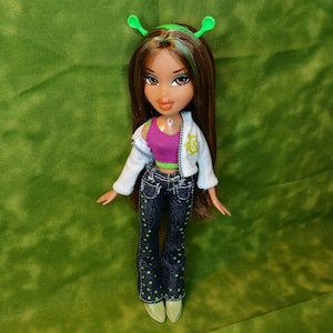 May include: A fashion doll with long brown hair with green streaks wearing a white jacket, a pink and green top, blue jeans with green polka dots, and a green headband with antennas.