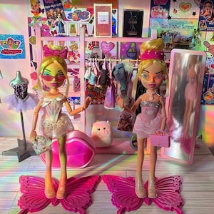 Puede incluir: Dos muñecas de moda con cabello rubio y gafas de sol están de pie sobre plataformas rosas con forma de mariposa. Están rodeadas de un armario rosa, accesorios y un espejo. Las muñecas visten atuendos elegantes, incluyendo vestidos y tacones.