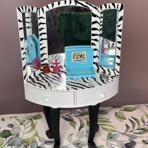Könnte beinhalten: Ein weiß-schwarzer Schminktisch mit Zebra-Streifen und einem dreifachen Spiegel. Der Schminktisch hat zwei Schubladen und schwarze Beine. Zubehör umfasst einen blauen Laptop, Parfümflaschen und ein kleines blaues Telefon. Der Schminktisch steht auf einer Oberfläche mit floralem Muster.