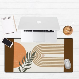 May include: A brown and white desk mat with a geometric design featuring a rainbow arch, a sun, and a leaf. The mat is on a white wooden surface with a laptop, a notebook, a pencil, and a cup of coffee.
