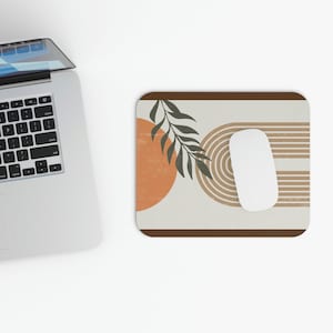 May include: A beige mouse pad with a brown border features an abstract design with an orange circle, brown stripes, and a green leaf.