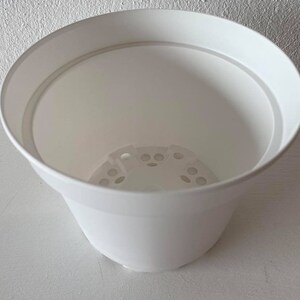 Op de afbeelding: Een witte plastic plantenpot met drainagegaten in de bodem. De pot is leeg en klaar om te planten.
