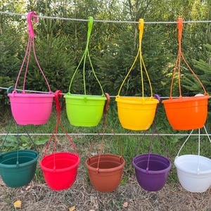 May include: A collection of nine round plastic hanging planters in various vibrant colours including pink, lime green, yellow, orange, dark green, red, brown, purple, and white, each featuring a matching plastic hanger attached to the rim.