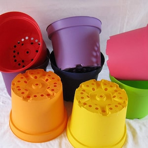 May include: A selection of brightly coloured plastic plant pots in shades of red, purple, pink, green, orange, and yellow. The pots are empty and have drainage holes. The pots are stacked on a white surface.