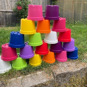 May include: A pyramid stack of twenty-one plastic plant pots in various vibrant colours including pink, purple, yellow, orange, red, blue, green, and white. Each pot features a wide rim and a base with drainage holes visible on the top.