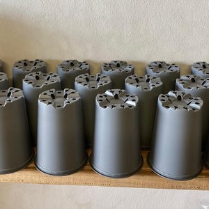 May include: A collection of twenty-one identical grey plastic plant pots arranged in rows. Each pot features a smooth, tapered cylindrical body and a base with multiple drainage holes and notched edges for ventilation and water flow.