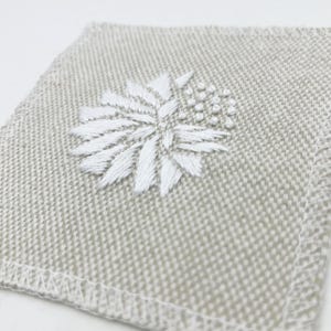 May include: A white embroidered flower design on a beige linen fabric coaster.