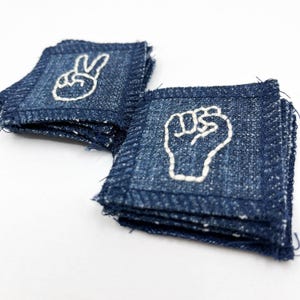 May include: Two denim patches with white embroidered designs. One patch features a peace sign, and the other displays a raised fist. The patches are square-shaped with frayed edges, and are stacked on a white surface.