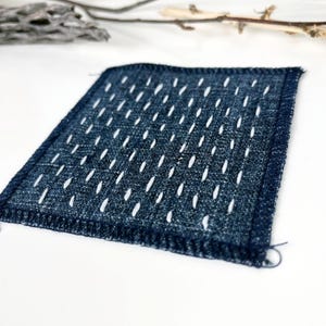 May include: A square, indigo-colored textile with a repeating pattern of white, elongated stitches. The edges are neatly finished with a dark blue border. The fabric has a textured appearance, suggesting a handmade or artisanal quality.
