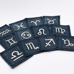 May include: A collection of denim patches embroidered with white thread, each displaying a different zodiac symbol. The patches are square-shaped and have a frayed edge, with symbols including Aries, Taurus, Gemini, and others.