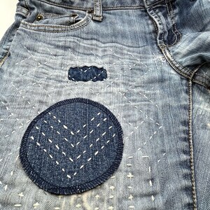 May include: Close-up of a pair of blue denim jeans with a circular patch sewn on the front. The patch is made of a darker blue denim and has white stitching in a geometric pattern.