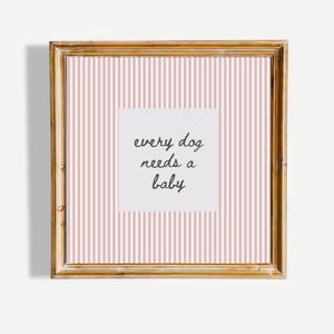Op de afbeelding: Een vierkant wandkunstwerk in een houten lijst met een lichtroze en witte verticaal gestreepte achtergrond. In het midden bevat een wit vierkant de zwarte cursieve tekst: every dog needs a baby, gecentreerd op de afdruk.