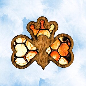 May include: A wooden brooch shaped like a bee with a crown on its head. The wings and crown are filled with a translucent, amber-colored resin featuring a dark honeycomb pattern. The body and frame are made of dark brown wood with a smooth finish.
