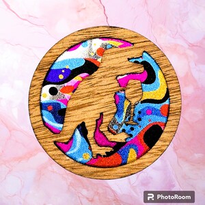 May include: A wooden round coaster with a cutout of a dinosaur silhouette. The dinosaur is filled with a colorful abstract design featuring swirls of pink, blue, yellow, and black.