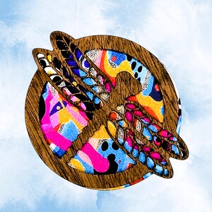 May include: A circular wooden brooch featuring a cutout dragonfly silhouette layered over a vibrant, multicolored background. The dragonfly has intricate wing details, while the background displays a mix of bright pink, yellow, and blue.