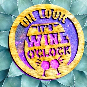 May include: Wooden coaster with a purple and pink glitter design. It says "It's Wine O'Clock" with two wine glasses.