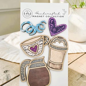 May include: A set of five wooden magnets on a white display card labeled Handcrafted MAGNET COLLECTION. The magnets include a blue swirl, a purple heart, a coffee cup with a heart, a tall coffee drink, and a coffee carafe with brown wood grain.