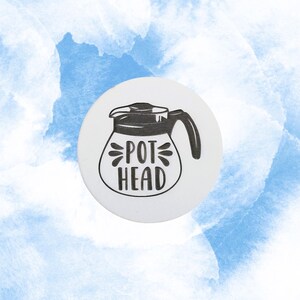 May include: A white, circular sticker featuring a black line drawing of a coffee pot with the words "POT HEAD" in a bold, stylized font. The background is a watercolor wash of blue and white.