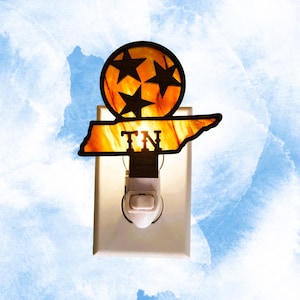 May include: A plug-in night light featuring a glowing amber-colored Tennessee state outline and a circular emblem with three black stars. The state shape includes the letters TN in black, and the entire piece is mounted on a white wall outlet.