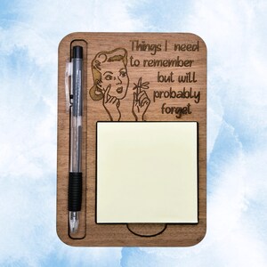 May include: A rectangular wooden note holder featuring a laser-engraved illustration of a person with a finger to their temple and the text "Things I need to remember but will probably forget." It includes a black pen and a square yellow sticky note.