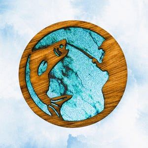 May include: A circular wooden coaster featuring a cutout silhouette of a jumping fish. The interior space is filled with a vibrant, shimmering turquoise resin that has a glittery, textured appearance against the natural brown wood grain.