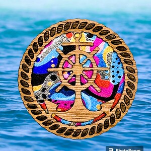 May include: A wooden nautical themed coaster with a colorful abstract design featuring a ship's wheel and anchor. The coaster is surrounded by a rope design.