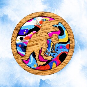 May include: A round wooden coaster with a colorful abstract design. The design features a mix of blue, pink, yellow, and black shapes against a light wood grain background. The coaster is set against a blue sky background.