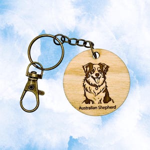 May include: A wooden round keychain with a laser-etched image of an Australian Shepherd dog. The dog is black, brown, and white, with the words "Australian Shepherd" below. The keychain has a bronze-colored clasp and ring.