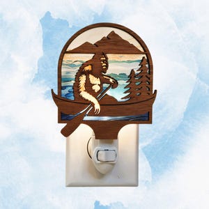 May include: A wooden night light featuring a Bigfoot figure paddling a canoe on a lake, with mountains and trees in the background. The night light is brown with a white outlet plug.