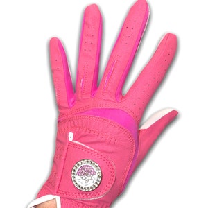 Op de afbeelding: Een felroze golfhandschoen met witte rand en een decoratief embleem. De handschoen is gemaakt van een glad materiaal met kleine perforaties voor ademend vermogen. Het embleem is een zilveren cirkel met een roze bloemenontwerp en heldere strass-steentjes.
