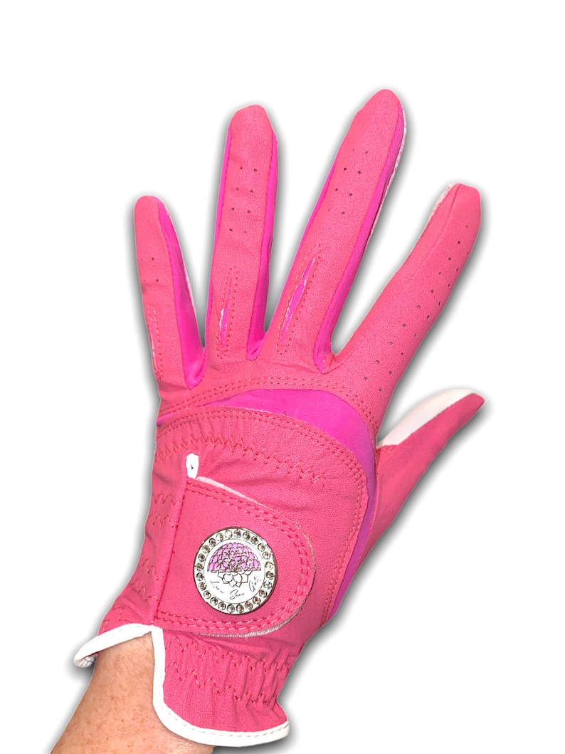 Op de afbeelding: Een felroze golfhandschoen met witte rand en een decoratief embleem. De handschoen is gemaakt van een glad materiaal met kleine perforaties voor ademend vermogen. Het embleem is een zilveren cirkel met een roze bloemenontwerp en heldere strass-steentjes.