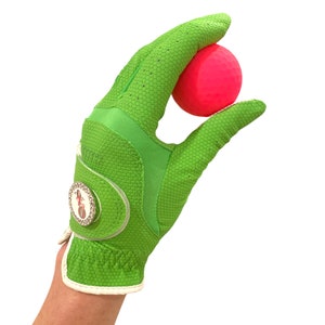 Op de afbeelding: Een felgroene golfhandschoen met een textuur houdt een felroze golfbal vast. De handschoen heeft een witte polsband en een decoratief embleem. De achtergrond is effen wit.
