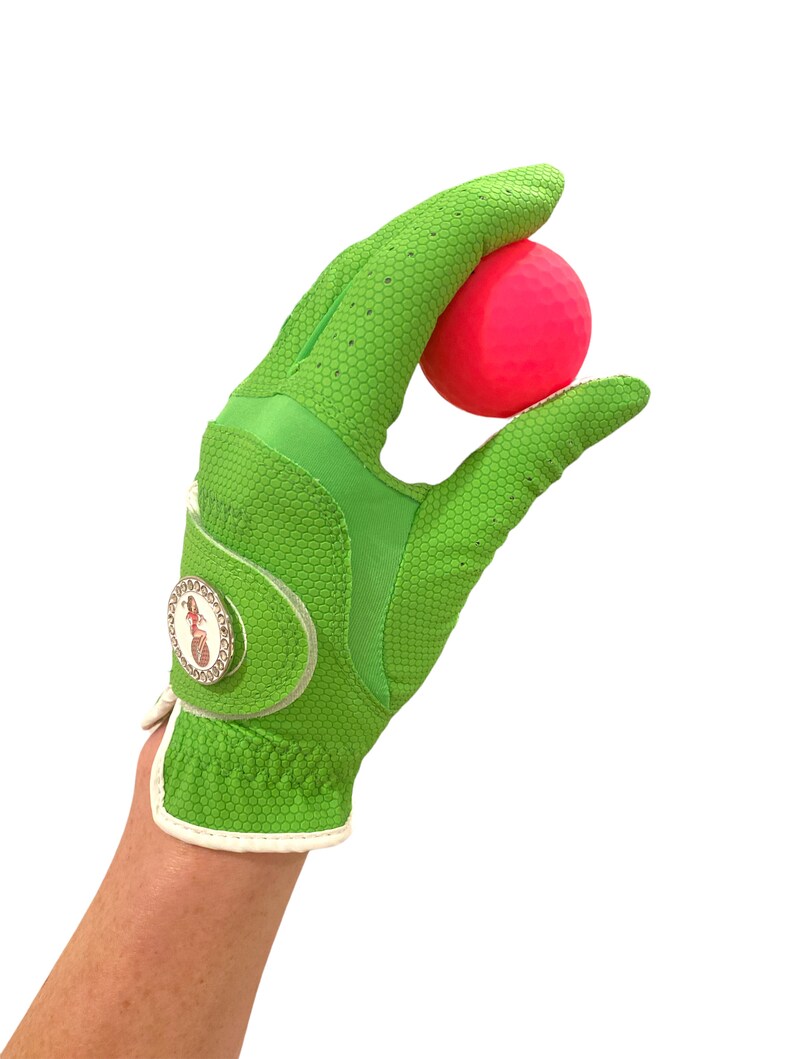 Op de afbeelding: Een felgroene golfhandschoen met een textuur houdt een felroze golfbal vast. De handschoen heeft een witte polsband en een decoratief embleem. De achtergrond is effen wit.