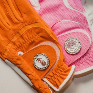 Puede incluir: Primer plano de dos guantes de golf, uno naranja y otro rosa. El guante naranja tiene un marcador de bola de golf decorativo con una mujer. El guante rosa tiene un marcador de bola de golf con un diseño floral. Ambos guantes tienen detalles en blanco.