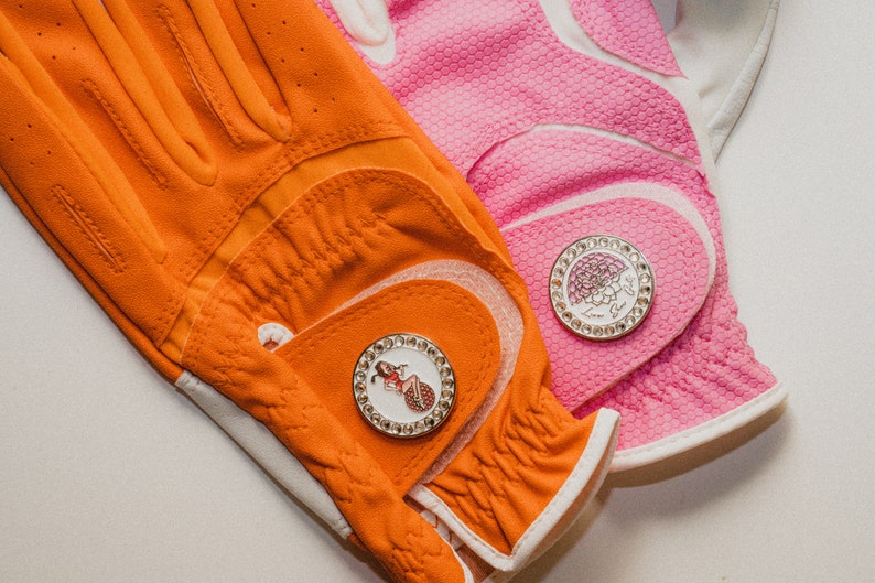Op de afbeelding: Close-up van twee golfhandschoenen, &eacute;&eacute;n oranje en &eacute;&eacute;n roze. De oranje handschoen heeft een decoratieve golfbalmarker met een vrouw erop. De roze handschoen heeft een golfbalmarker met een bloemdesign. Beide handschoenen hebben witte accenten.