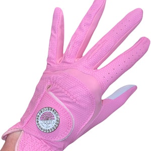 Op de afbeelding: Een roze golfhandschoen met een honingraattextuur. De handschoen heeft een wit accent op de palm en een cirkelvormig embleem met een bloemenontwerp en kristallen details. De handschoen is ontworpen voor golf.