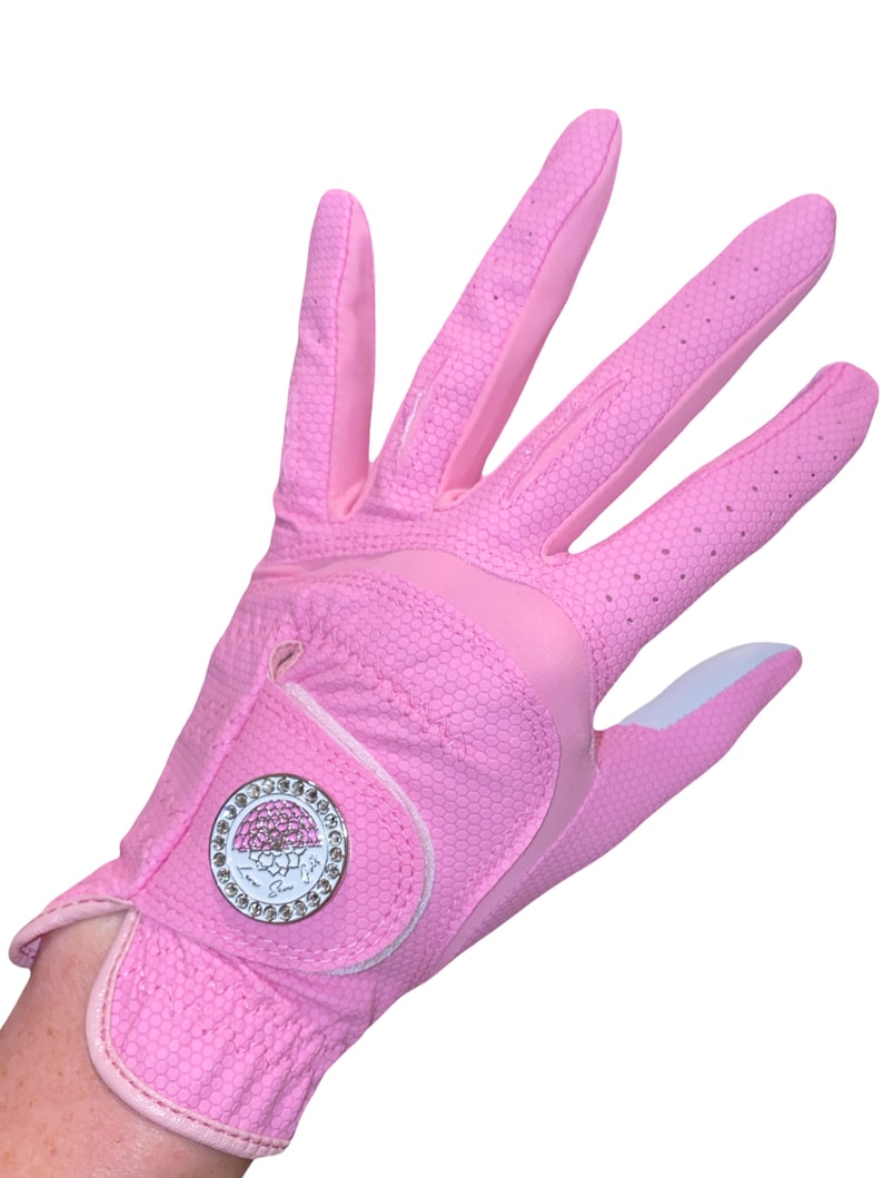 Op de afbeelding: Een roze golfhandschoen met een honingraattextuur. De handschoen heeft een wit accent op de palm en een cirkelvormig embleem met een bloemenontwerp en kristallen details. De handschoen is ontworpen voor golf.